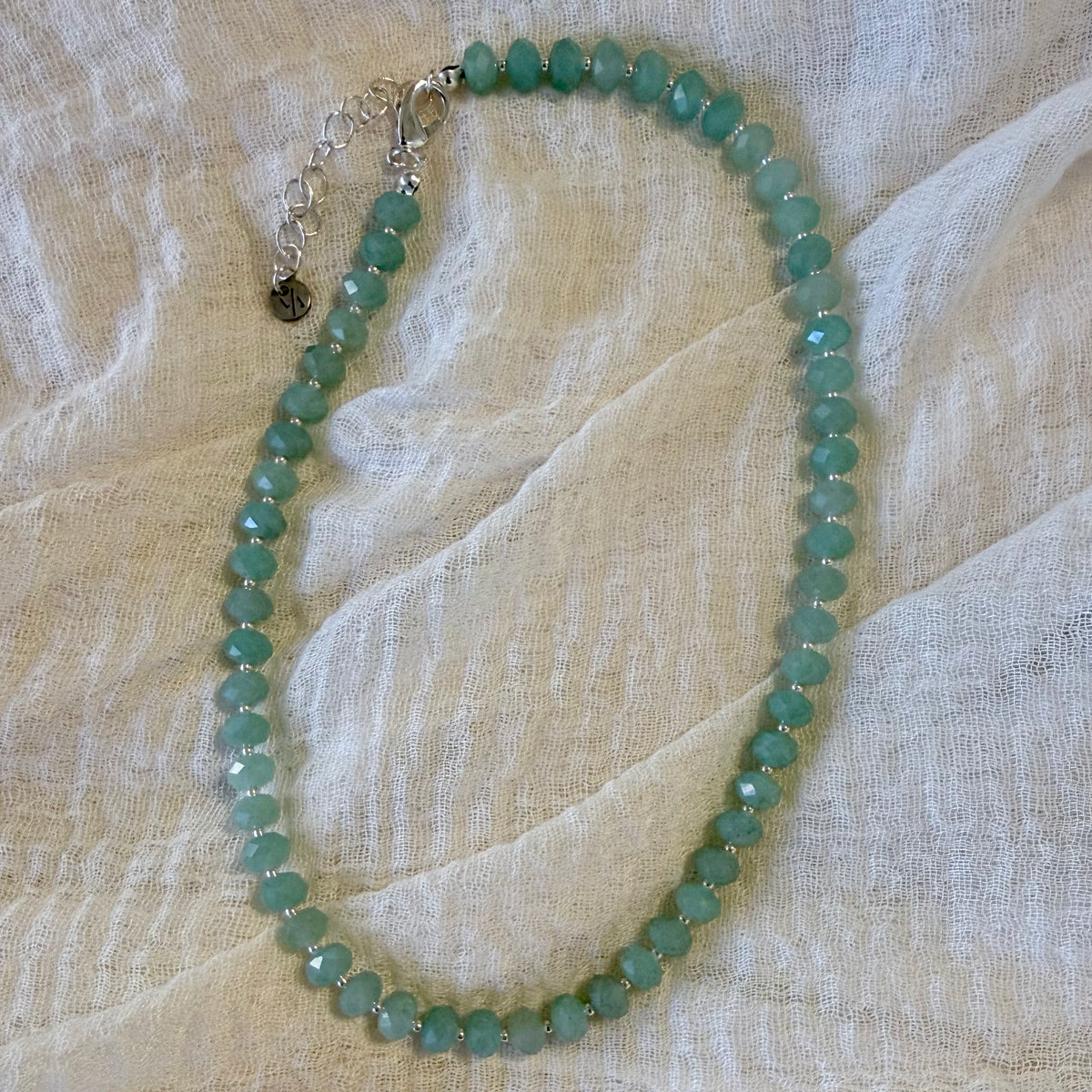A beaded necklace made of faceted light green beads with a silver-tone clasp, laid flat on a textured off-white fabric background.