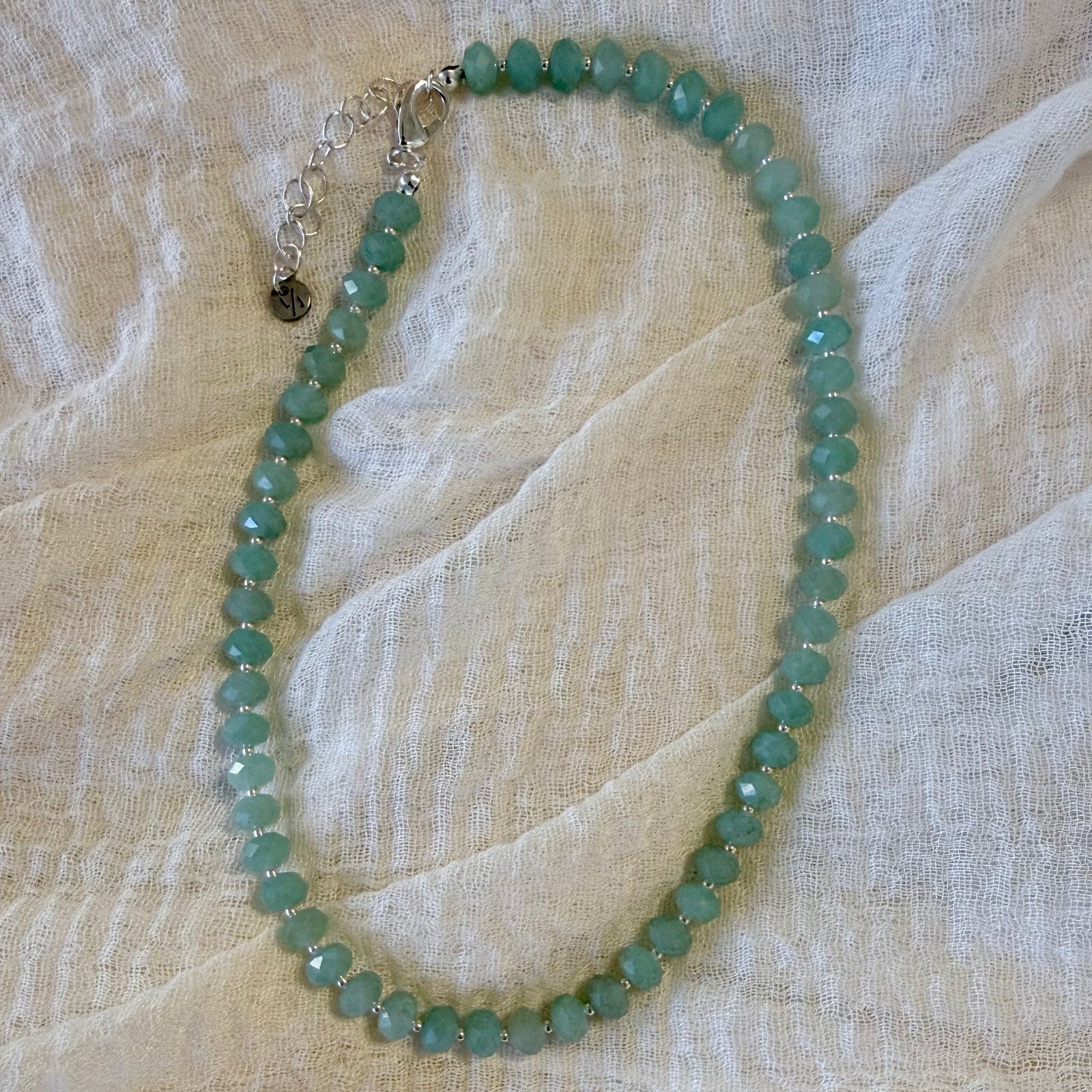 Faceted light green jade bead necklace with small silver spacer beads, draped on a textured cream-colored fabric.