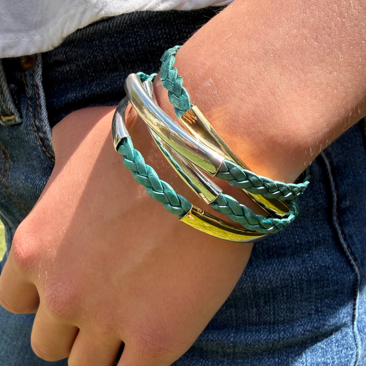 A close-up of a person's wrist wearing a stack of multi-layered bracelets, including braided mint green leather and polished silver and gold-tone metal bands, paired with blue denim jeans.