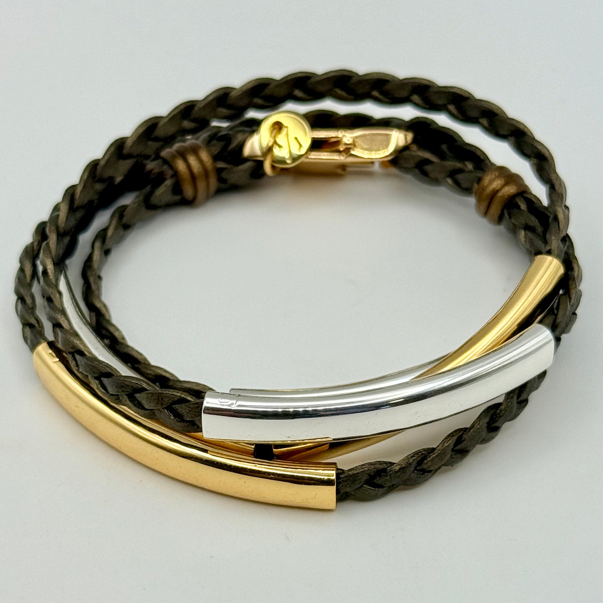 A multi-strand wrap bracelet featuring dark brown braided leather cords, polished gold-toned curved bar elements, a polished silver-toned bar, and a gold lobster clasp, shown on a light gray background.