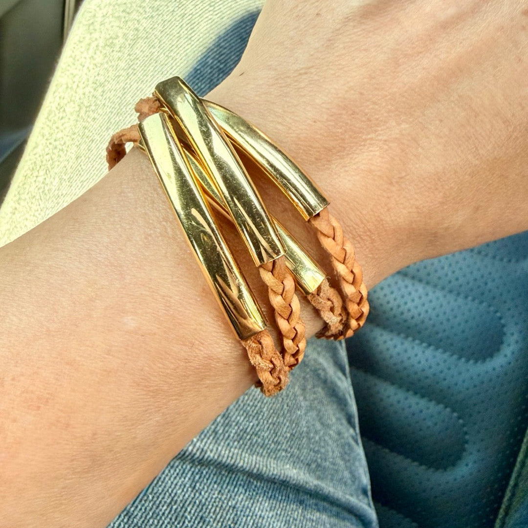 A close-up of a person's wrist wearing a stack of bracelets, including two polished gold metal tubes and two tan braided leather cords, with blue jeans and a car seat visible in the background.