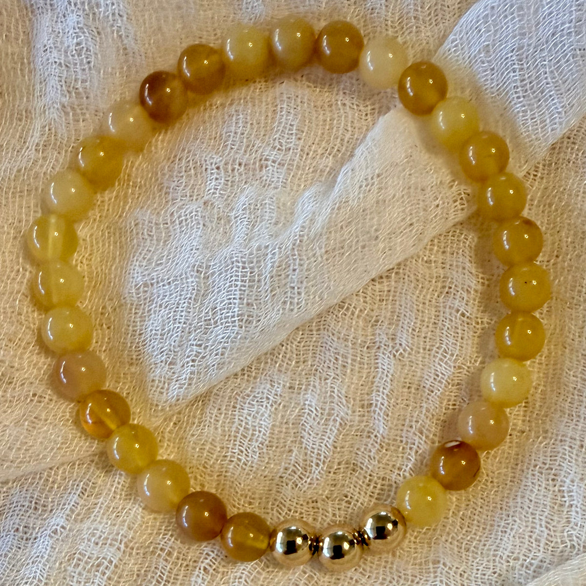 A beaded stretch bracelet made of translucent yellow and amber round beads, with three small metallic gold spacer beads at the bottom, displayed on a textured cream fabric background.
