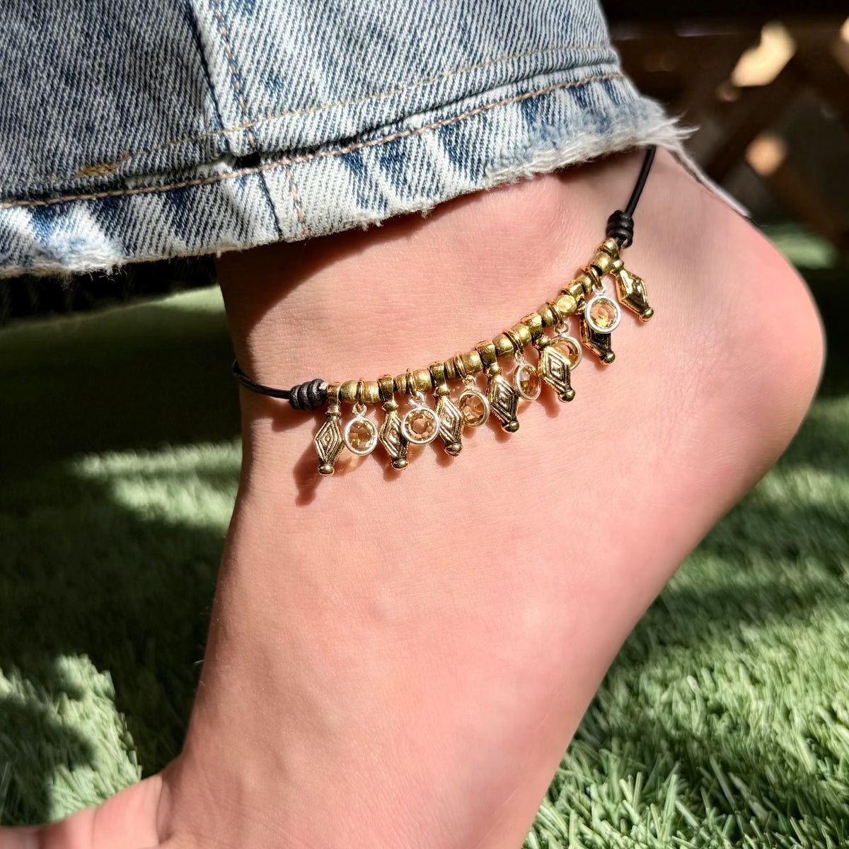 Ankle bracelet with gold charms on a person's ankle against a grassy background