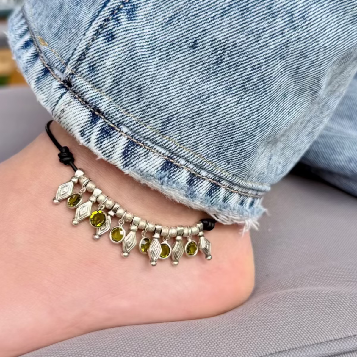 Anchored anklet with charms on a foot wearing blue jeans, sitting on a gray surface.