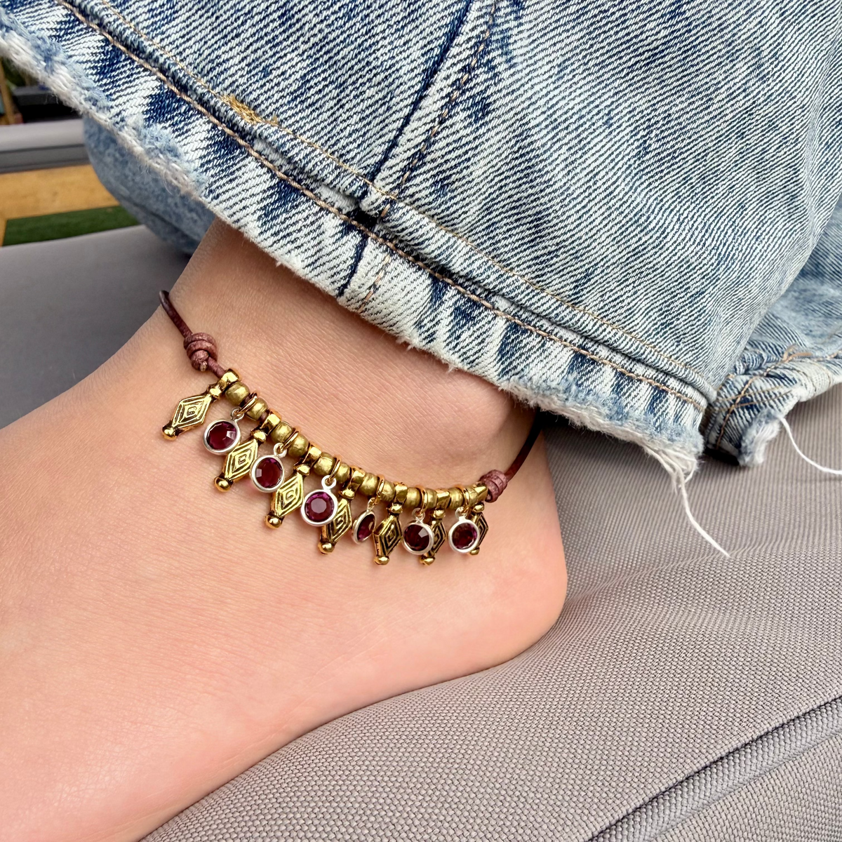 Ankle with a gold anklet featuring red gemstones, wearing denim jeans.