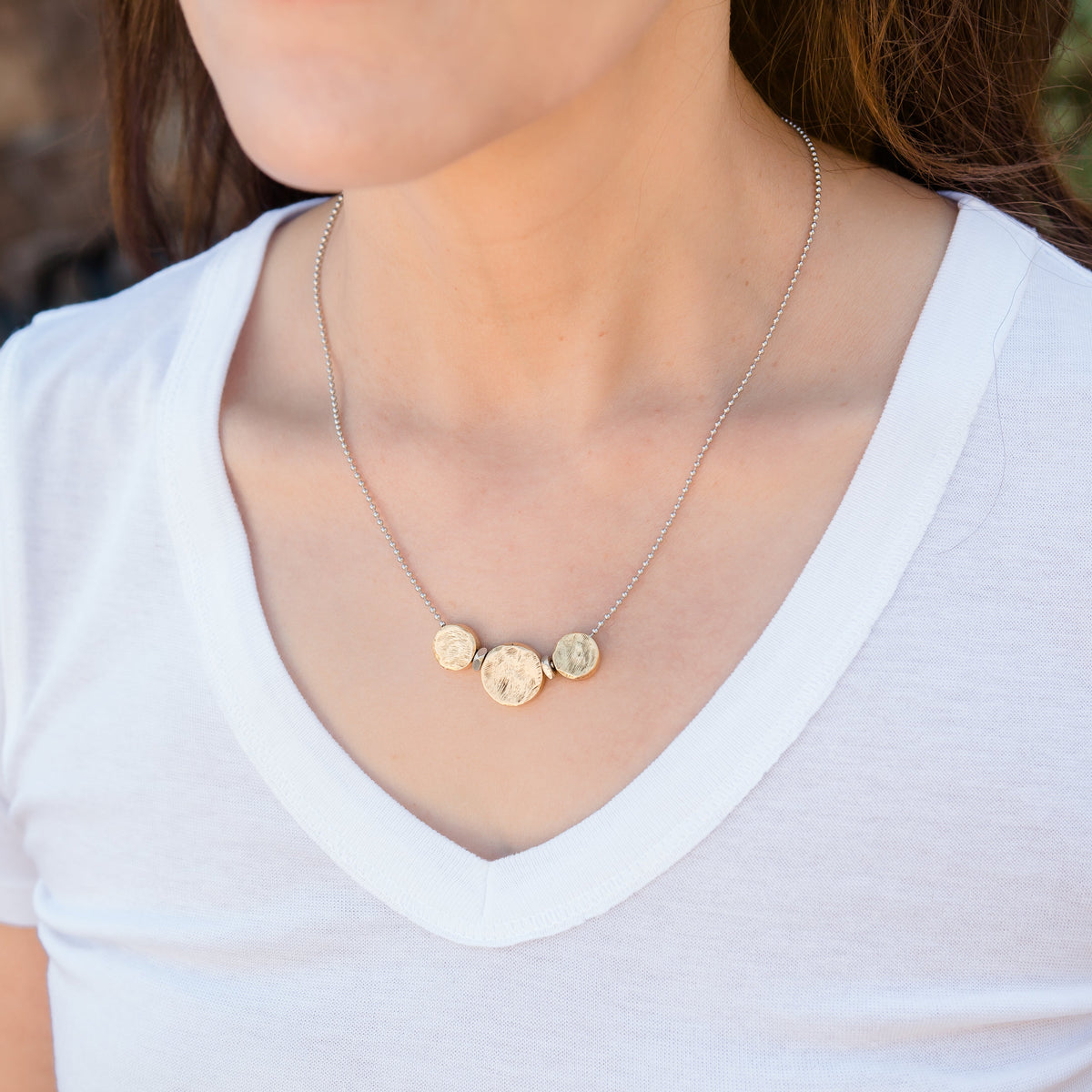 Person wearing a gold necklace with three circular pendants on a white shirt.