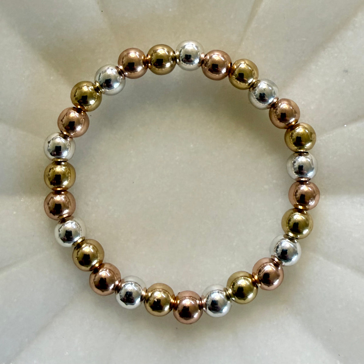 A beaded stretch bracelet made of alternating metallic gold, rose gold, and silver round beads, displayed on a textured white background.