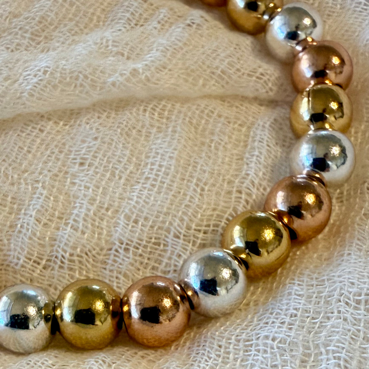 A close-up of a beaded necklace with alternating shiny metallic gold and silver beads, resting on a textured cream-colored fabric background.