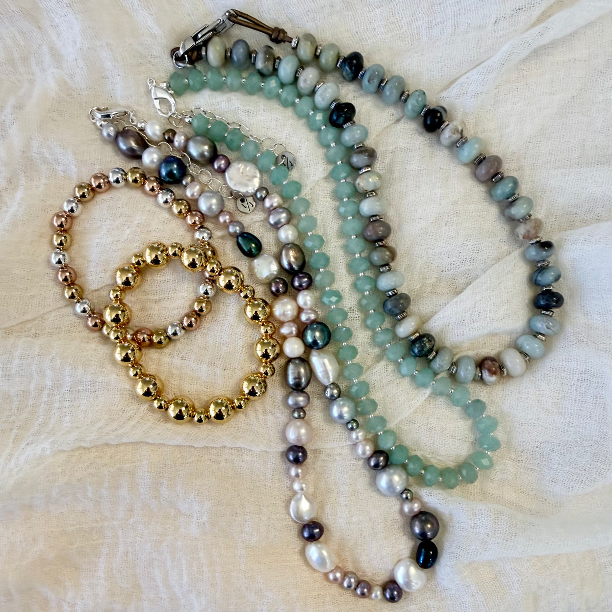 A collection of handmade beaded jewelry, including a long necklace and two bracelets, arranged on a textured cream fabric background. The necklace features light green jade-like beads, gray and black pearls, and white pearls, while the bracelets are made of gold and silver-toned seed beads.