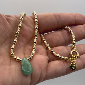 Video of hand holding Gold beaded necklace with a green teardrop pendant on a light gray background